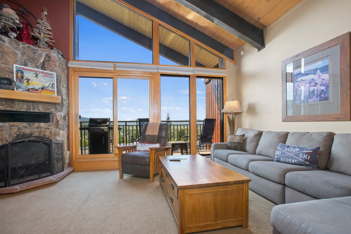 The image showcases a beautifully furnished condo living room with a striking stone fireplace as the centerpiece, surrounded by plush sofa seating and a rustic wooden coffee table. Large windows flood the space with natural light and offer stunning sunset mountain views, creating an inviting atmosphere perfect for gathering with friends or family. The overall vibe is cozy yet upscale, ideal for relaxing after a day on the slopes or enjoying a summer evening.