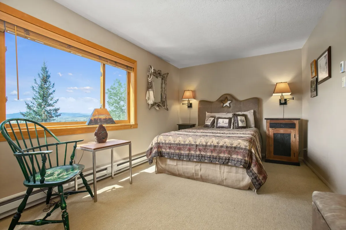 A serene bedroom featuring a bed with patterned bedding, a large window with scenic mountain views, a green chair for relaxation, and a bedside lamp. The space exudes comfort and tranquility, ideal for unwinding after a day on the slopes.