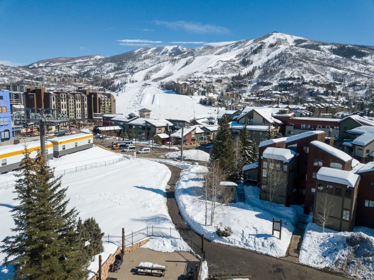 A scenic panorama of the Steamboat base area featuring snow-topped mountains, a busy ski lift, town buildings, and a winding pathway that beckons exploration. The blend of natural beauty and bustling village life is palpable.