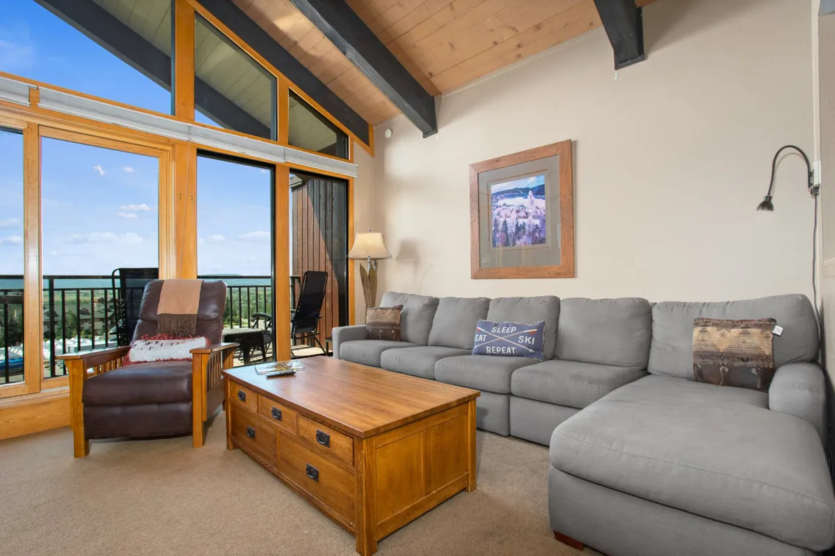 A spacious living room featuring a large sectional sofa, wooden coffee table, and an armchair placed near expansive windows. Framed artwork adorns the wall, and the room is bathed in natural light, with cozy furnishings arranged for social gatherings and relaxation. The view through the windows hints at the snowy slopes outside, tying the interior to its mountain setting.