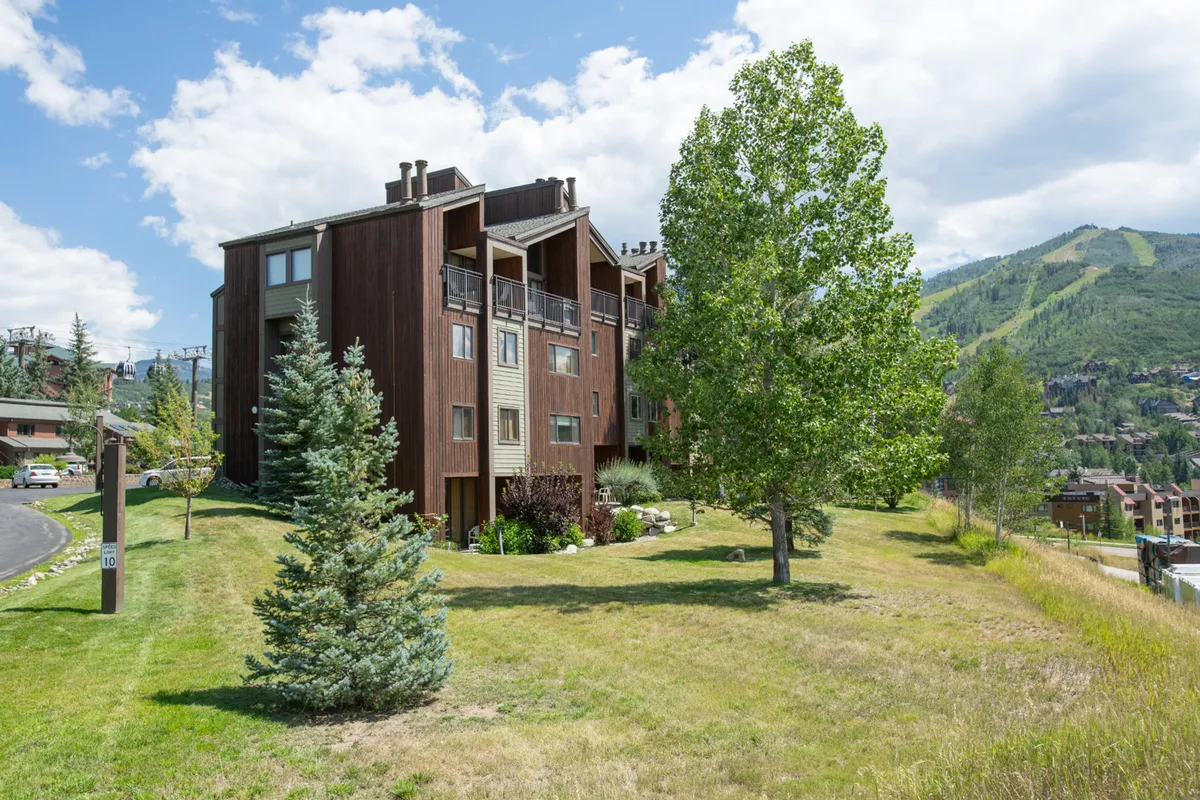 A wide-angle view of the property's exterior with lush summer greenery, set against a backdrop of surrounding mountains. The scene suggests easy access to hiking trails and outdoor gathering areas, capturing the inviting natural environment in warm weather.