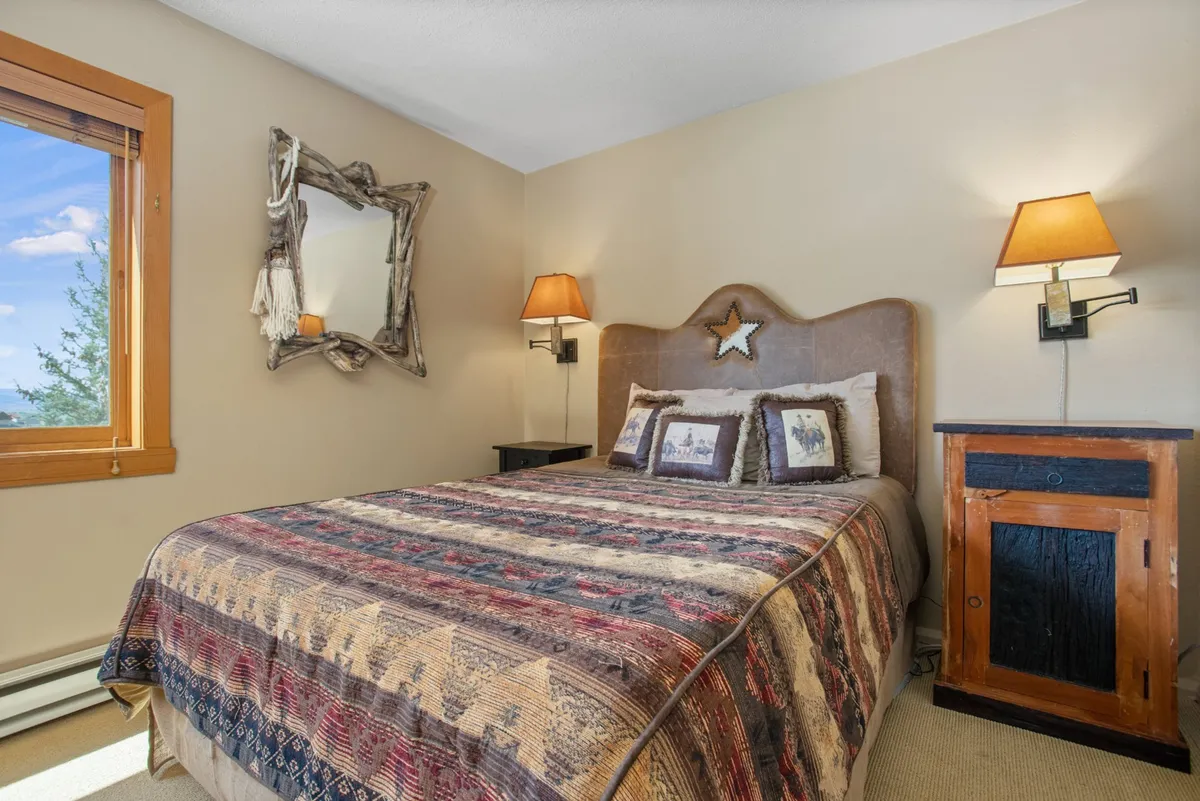 An inviting bedroom with a bed dressed in a patterned blanket and decorative pillows, flanked by wooden nightstands and soft bedside lamps. A wooden mirror adds to the warmth and charm, and the lighting is soft and restful.