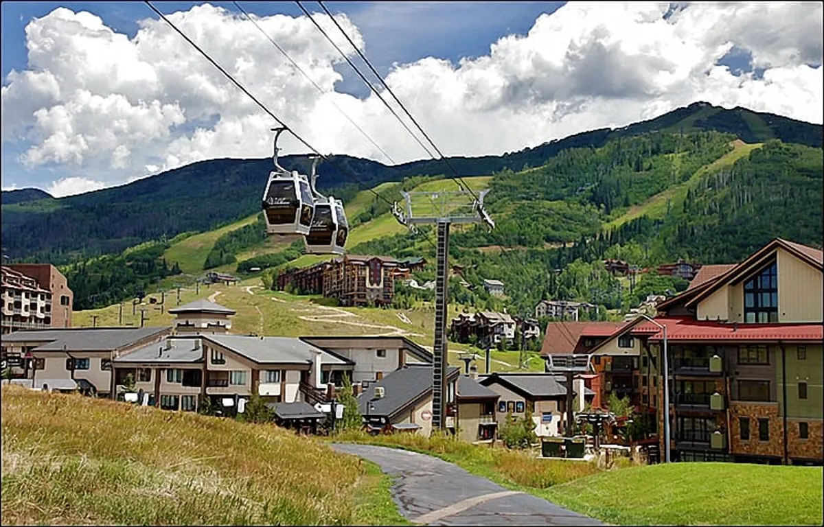 This exterior image captures the ski resort buildings with the gondola lift prominently featured against a stunning mountain backdrop. The hillside is lush and green, indicating proximity to both mountain activities and the gondola terminal, while the architecture and landscape exude the excitement and accessibility of a ski-in/ski-out destination.