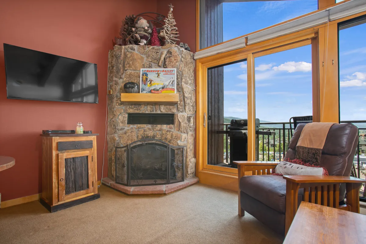 A welcoming interior with a striking stone fireplace as the focal point, complemented by wooden furniture and a television. Large windows offer glimpses of the outdoors, while the seating area invites relaxation by the fire or gathering for a movie night.