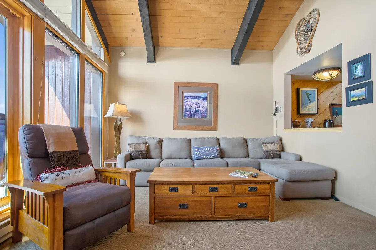 A bright, relaxed living space featuring a comfortable seating arrangement around a sturdy wooden coffee table. Large windows welcome abundant natural light and showcase the snowy exterior, while the setting is ideal for socializing, game nights, or simply enjoying the mountain views.