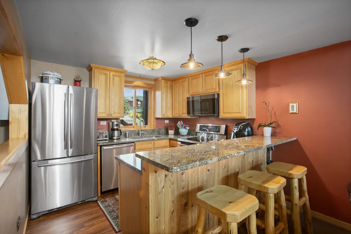 A warm and inviting kitchen featuring a granite countertop, wooden bar stools, modern stainless steel appliances, and a window that offers a view of the outdoors. The space feels open and ready for both cooking and socializing, with plenty of room for preparing meals and hosting guests.