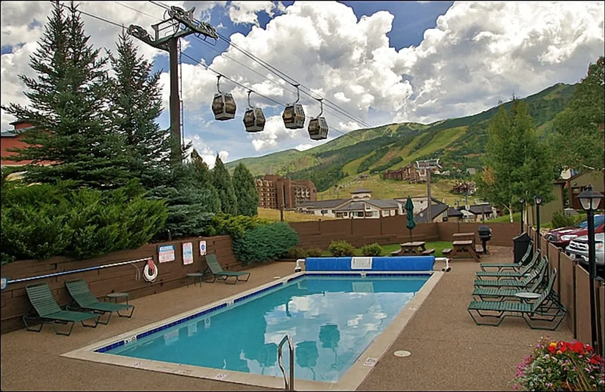 A vibrant resort pool area with inviting lounge chairs and a gondola lift passing overhead, set against a stunning mountain backdrop. The lively yet relaxed atmosphere hints at both fun and tranquility.