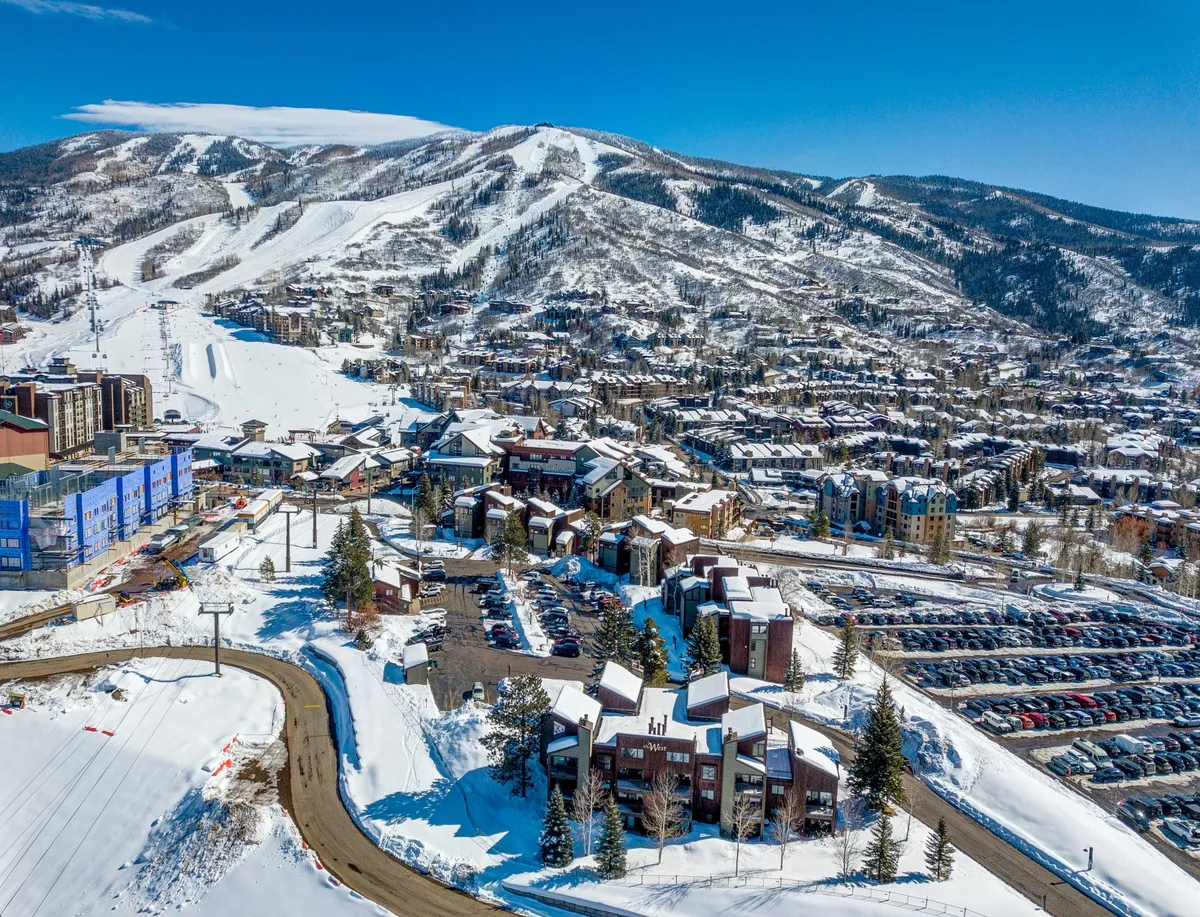 An inviting view of snow-covered slopes, residential buildings, and nearby restaurants nestled at the base of the mountains. Warm lights and cozy architecture suggest a welcoming après-ski environment.