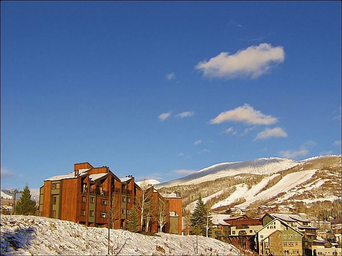 A scenic exterior shot showcasing a wooden lodge-style building set against the backdrop of snow-covered mountains and a clear blue sky. The view highlights the mountain surroundings and the inviting architecture of the property.