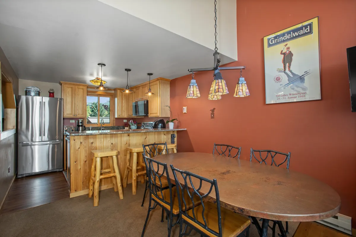 A rustic yet modern dining area with a wooden table set beneath colorful pendant lights. The kitchen cabinetry is visible, tying the dining and cooking spaces together, while a vintage Grindelwald poster adds character and warmth to the atmosphere.